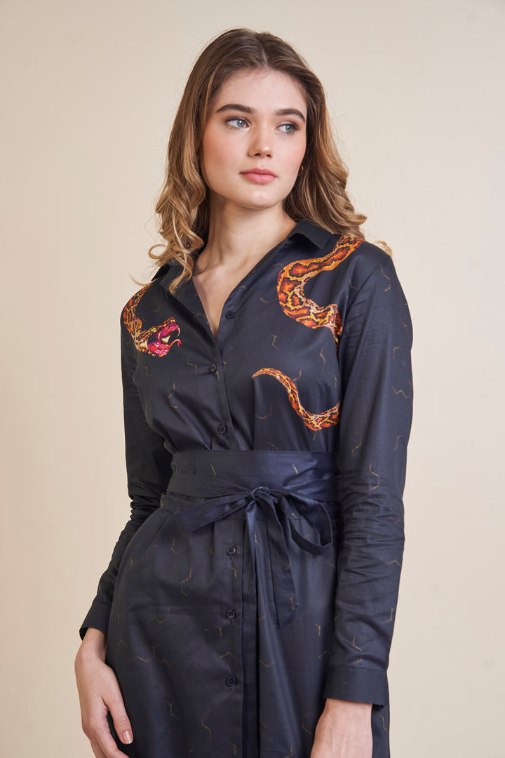 A woman standing in a snake print shirt dress, with roll-up sleeves and a belted waist. The dress features a collar and button-front design, making it a versatile and stylish choice for resortwear or everyday wear